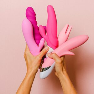 Hands holding various pink silicone vibrators against a pastel pink backdrop.