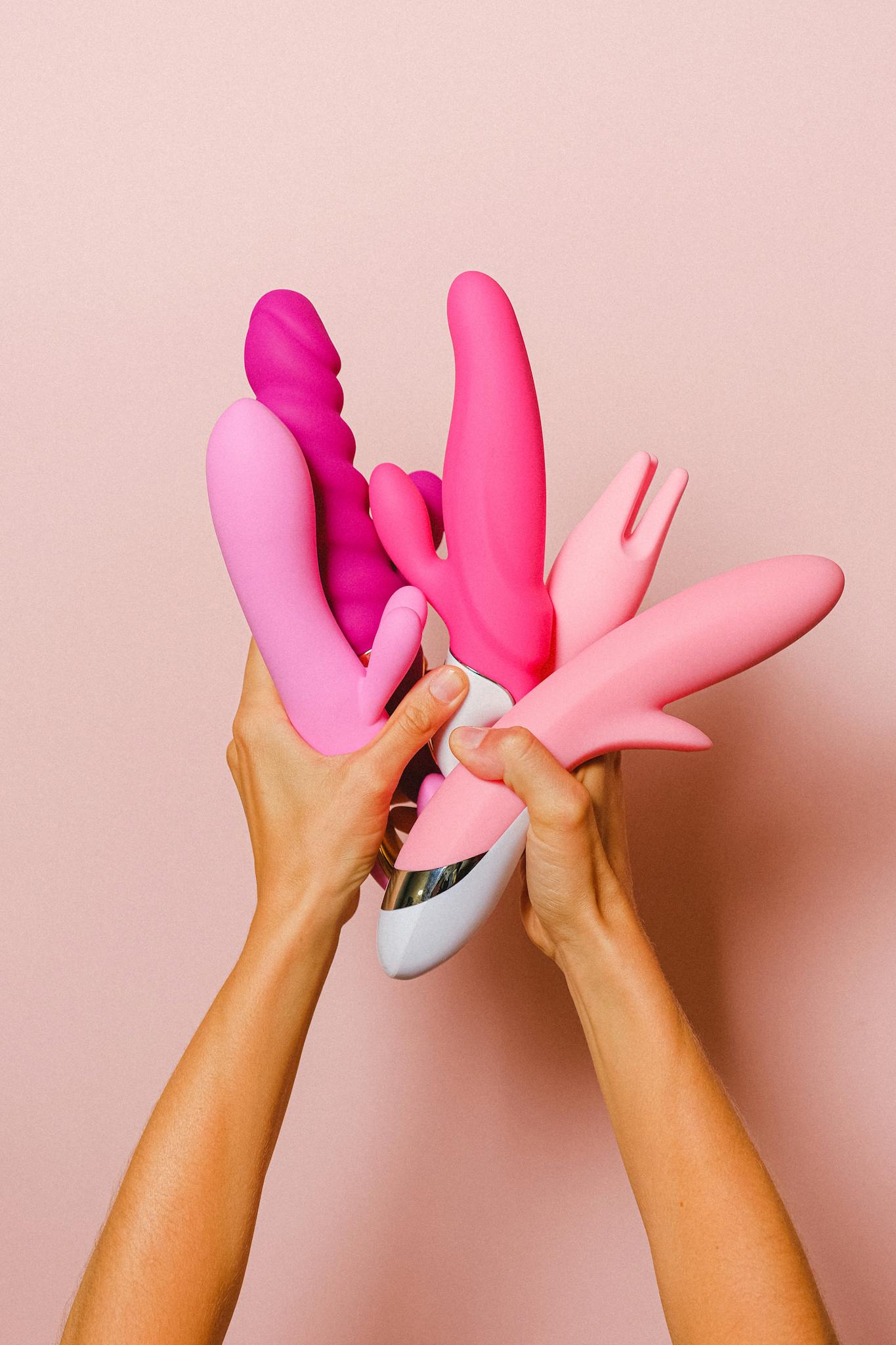 Hands holding various pink silicone vibrators against a pastel pink backdrop.