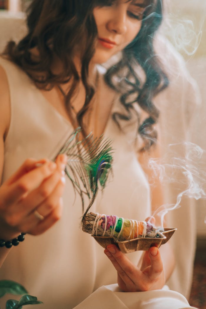 Crop female with wavy dark hair smiling and smelling smoke of smudge sticks on blurred background